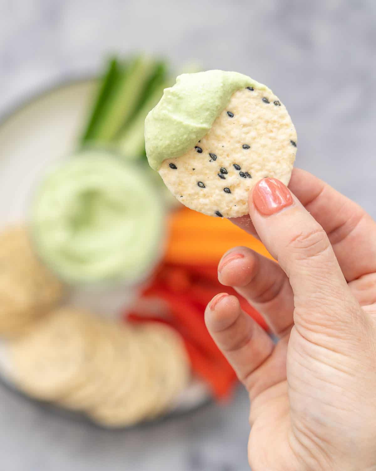 Cracker being held up to camera with cottage cheese dip on it. Cucumber, carrot and capsicum slices and crackers on a plate with a glass ramekin of cottage cheese dip blurred in background.