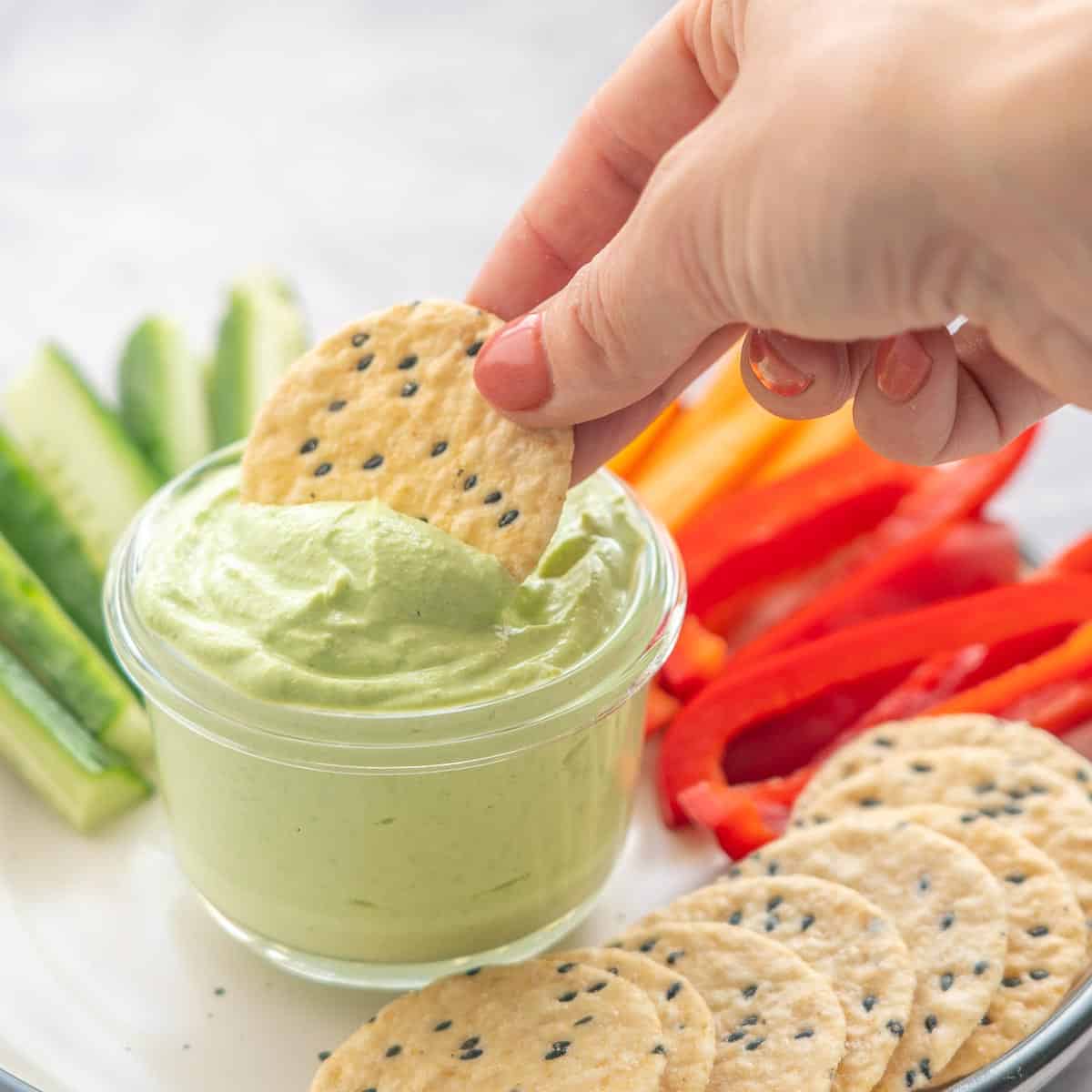 A plate full of crackers and sliced fresh capsicums, carrots and cucumbers with a hand dunking a cracker into a  ramekin of Cottage Cheese Dip 