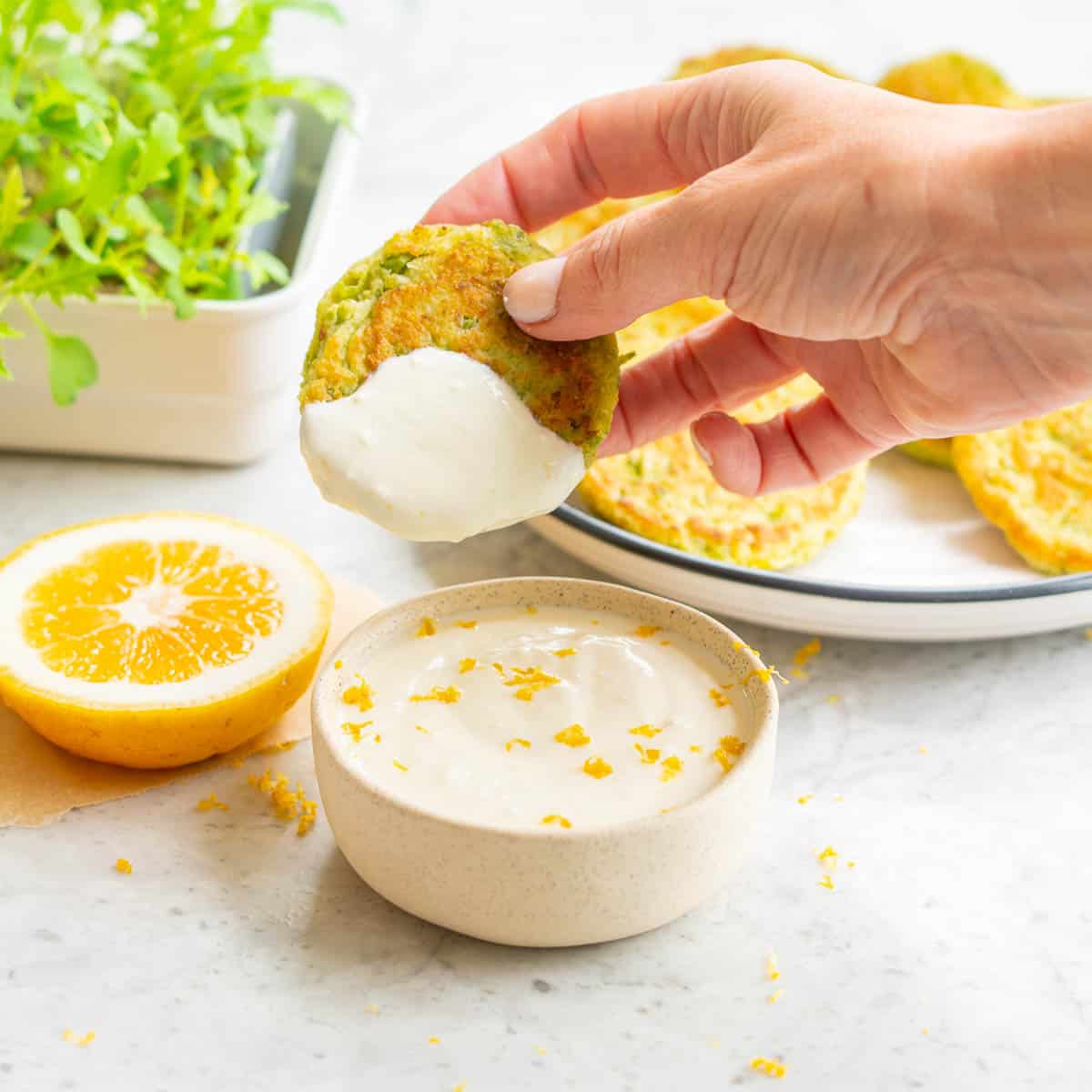 A hand holding a pea fritter which has been dipped into a yoghurt dressing which is sitting below next to a slice of lemon, a bowl full of green salad and a plate of pea fritters