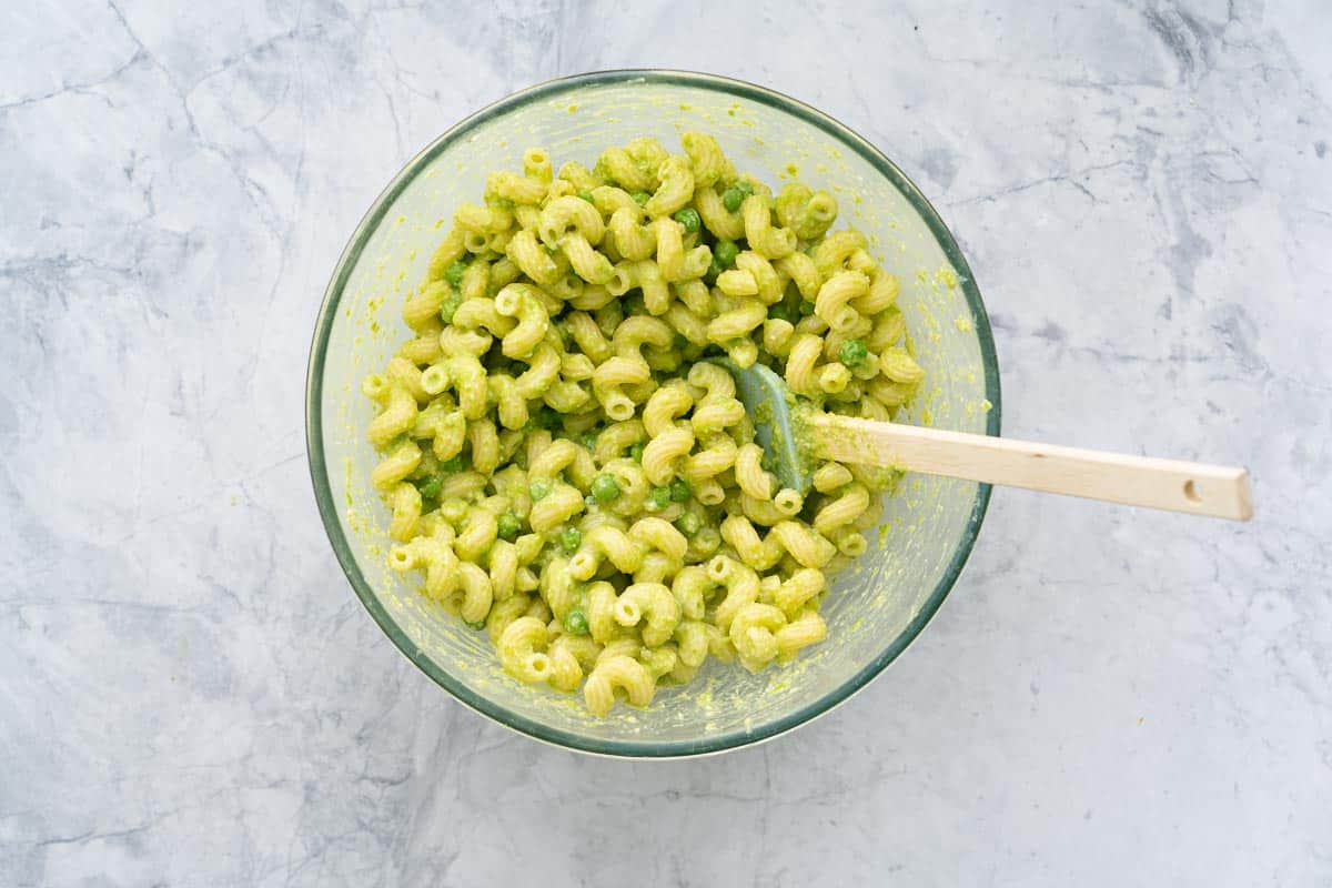 A large glass bowl of pea pasta sitting on a bench with a wooden spoon resting against the side.