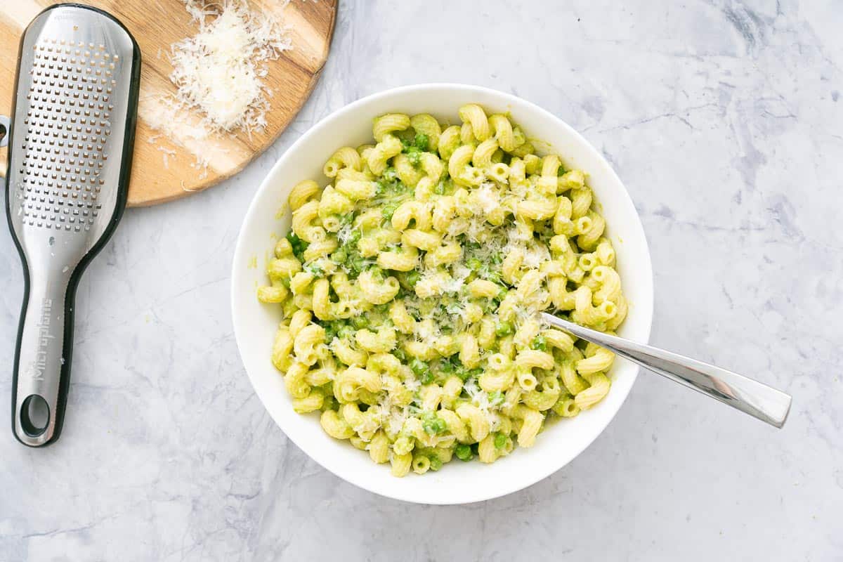 A large serving of Pea Pasta in a bowl sprinkled with parmesan cheese sitting on the bench with a spoon resting on the side. And a wooden serving board with a grater and sprinkled parmesan cheese.