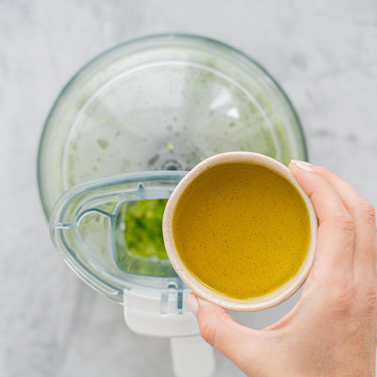A small ramekin of oil held by a hand above the food processor.