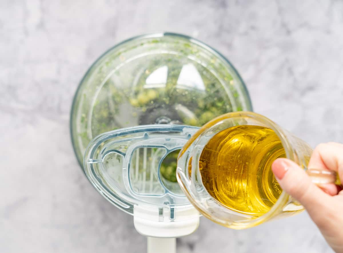 A glass jug of oil being poured in to the basil and spinach mixture through the top of the blender