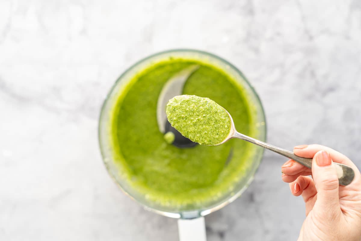 A spoon full of pesto being held above the mixture below in a food processor