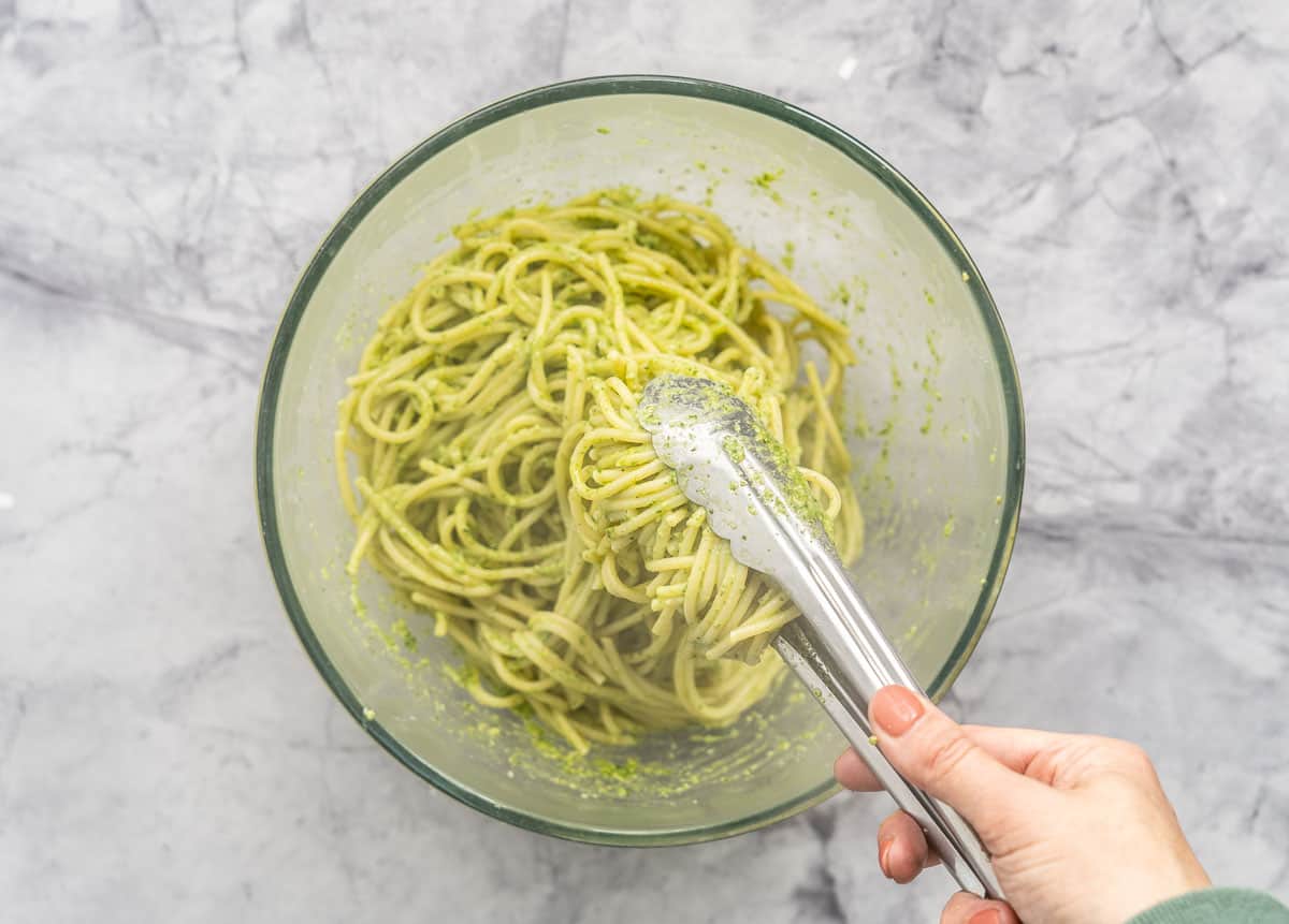A glass bowl full of tossed Pesto and Pasta sitting on the bench with a pair of tongs mixing it together
