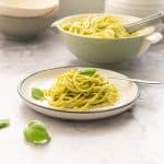 A plate of spaghetti coated in green pesto in front of a large bowl of pasta.