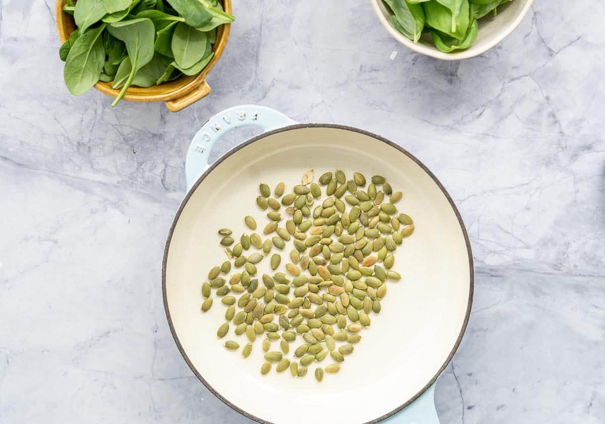 Toasted pepitas scattered along the bottom of a frying pan which is sitting on the bench next to a bowl eachof spinach and basil