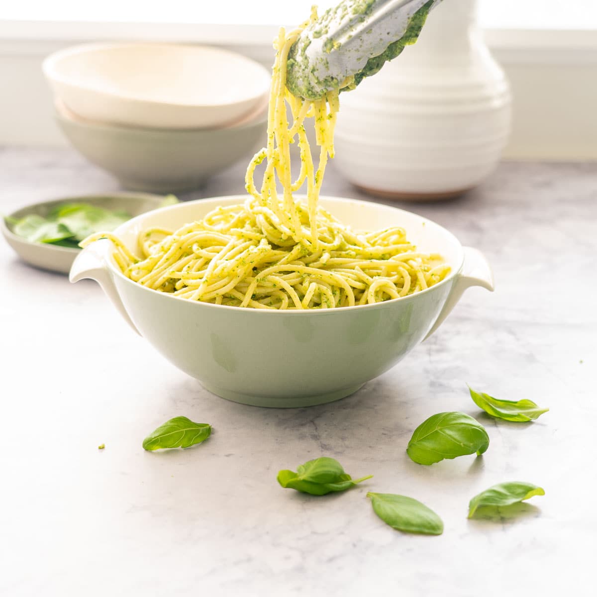 A large bowl filled with pesto pasta sitting on the bench with stacked bowls and a scattering of basil leaves around on the bench