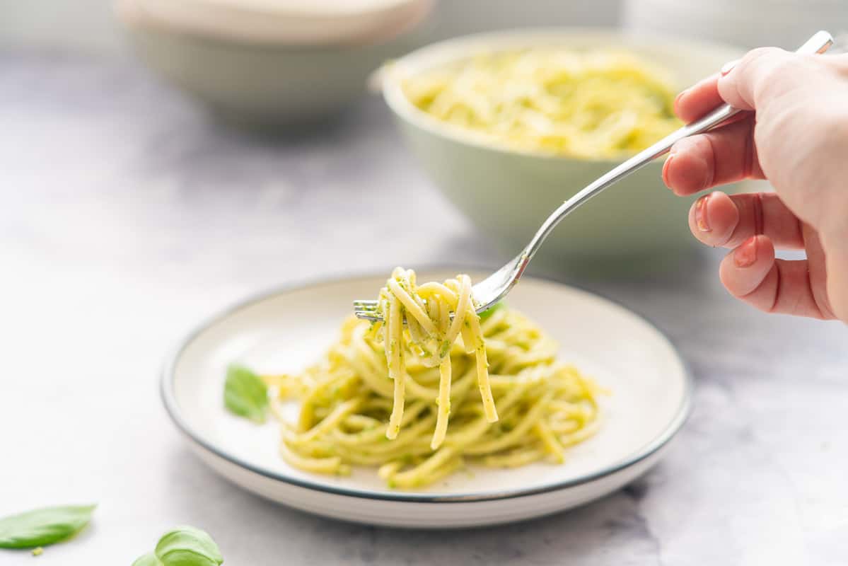 A plate full of pesto pasta with a hand holding a forkful of the pasta above it.