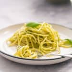 A plate full of pesto pasta with a hand holding a forkful of the pasta above it.