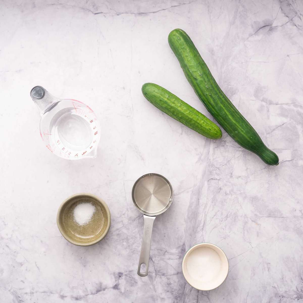 Cucumbers, a jug of water, vinegar, salt and sugar laid out on a marble bench top.