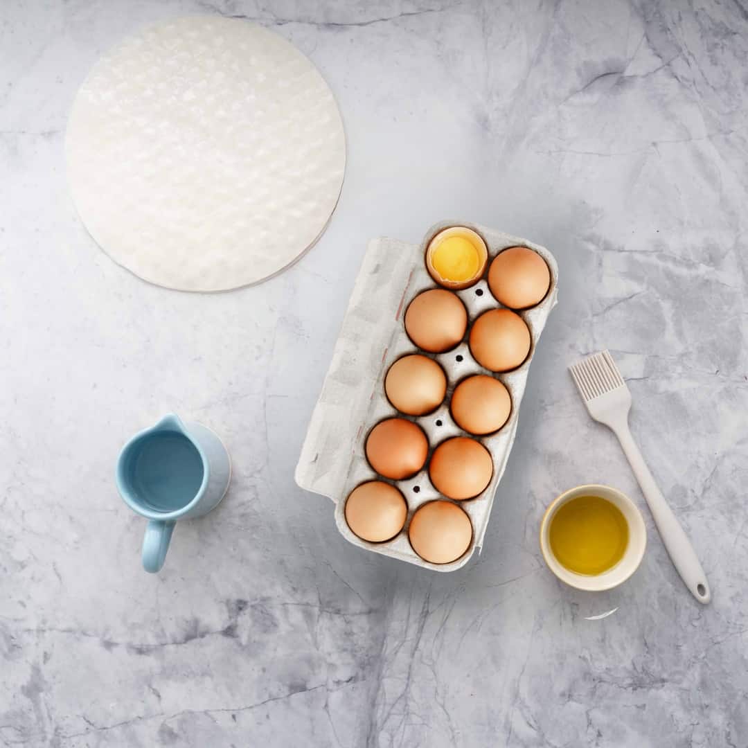 A dozen eggs, jug of water, bowl of oil and rice paper sheets on a marble bench top