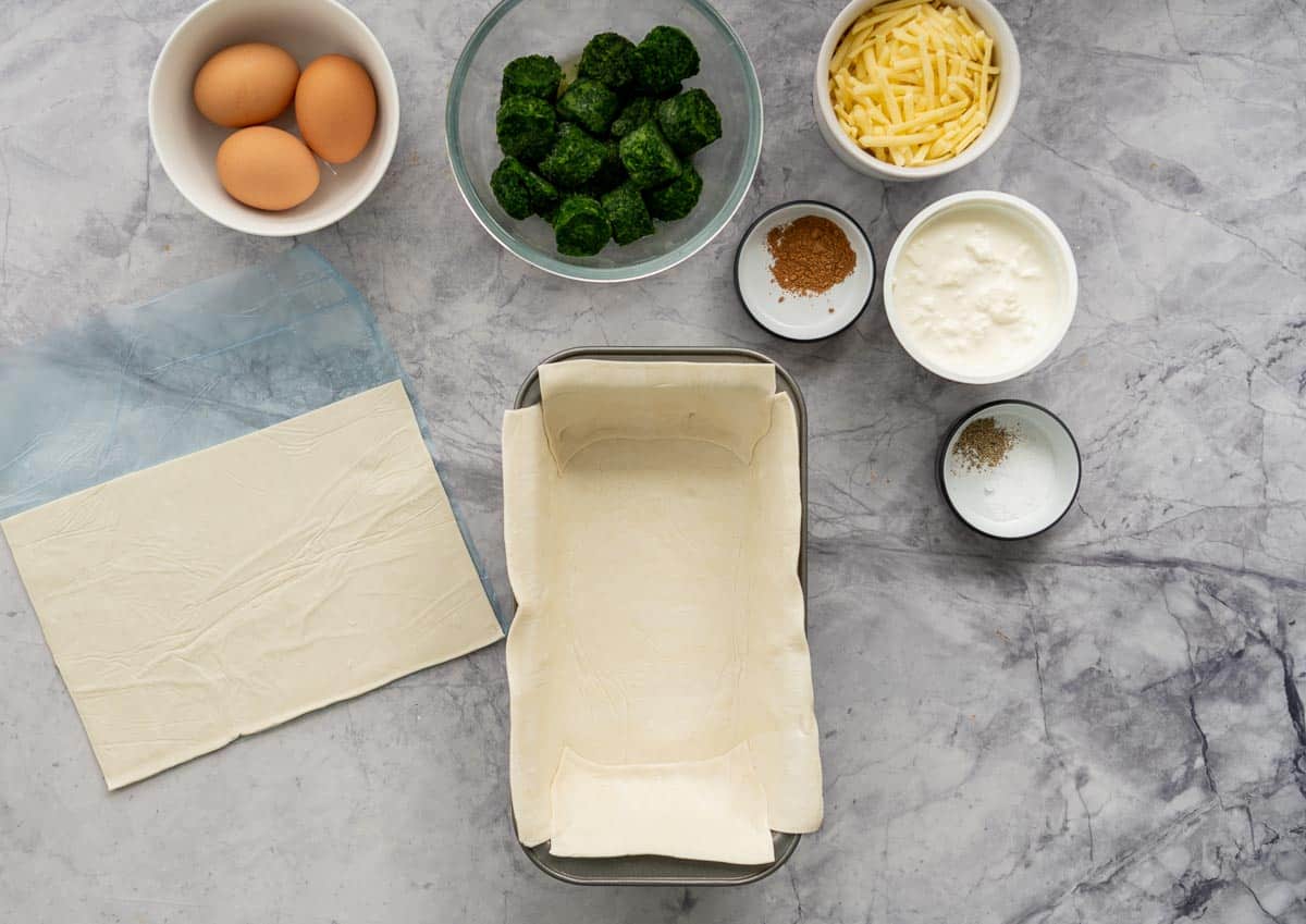 A loaf tin lined with pastry on a bench surrounded by other ingredients.