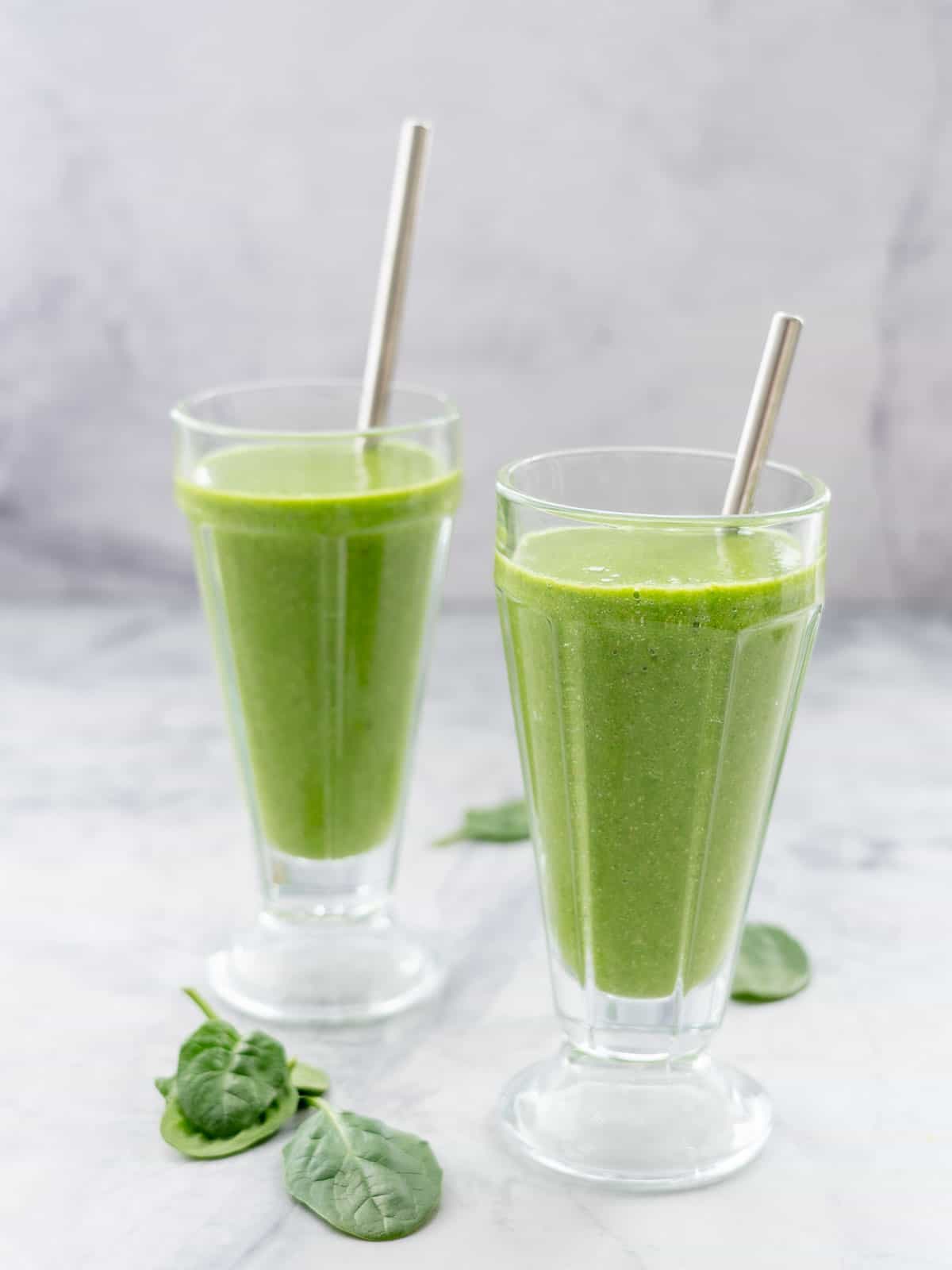 Two tall glasses with metal straws in each of them, filled with bright green smoothie sitting on a bench with a scattering of spinach leaves