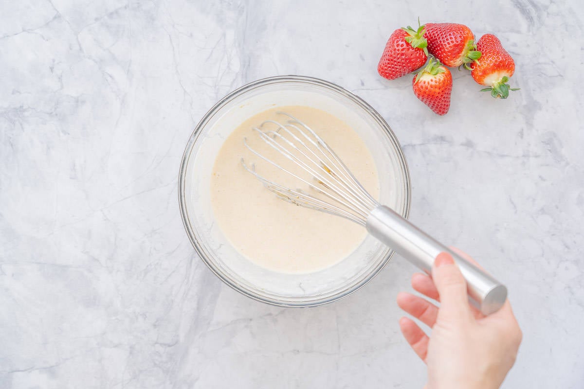 A large glass bowl with a hand whisking ingredients together with a handful of strawberries sitting on the bench to the right.