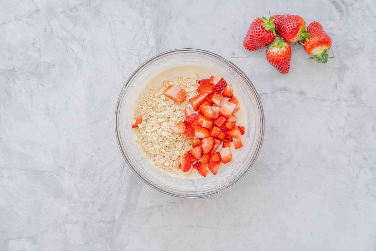 A large glass bowl sitting on the bench filled with ingredients to make the strawberry overnight oats sitting on the bench with a handful of strawberries sitting on the bench to the right