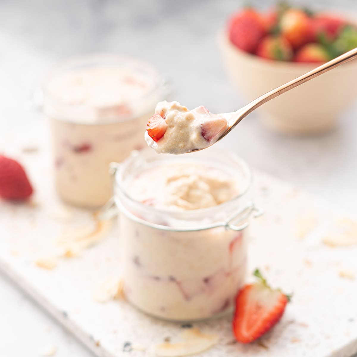 A spoonful of strawberry overnight oats held above 2 full jars sitting on a terrazzo serving board with a scattering of toasted coconut flakes and strawberry halves