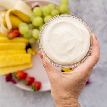 a glass bowl of whipped cottage cheese being held up to the camera above a fresh fruit platter.