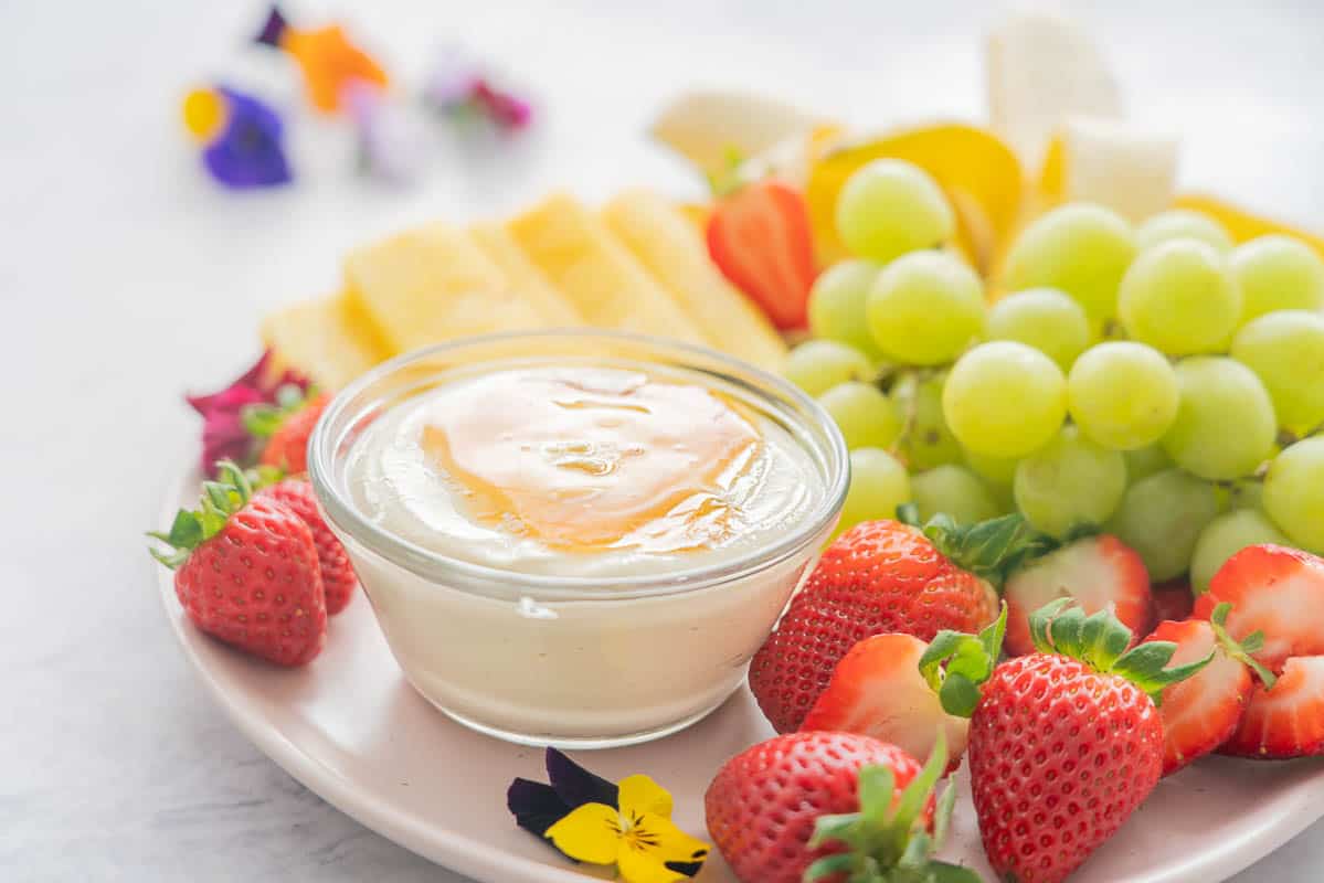 A plate full of fresh strawberries, grapes, bananas and pineapple and the Whipped cottage cheese dip in a glass ramekin with honey drizzled on the top.