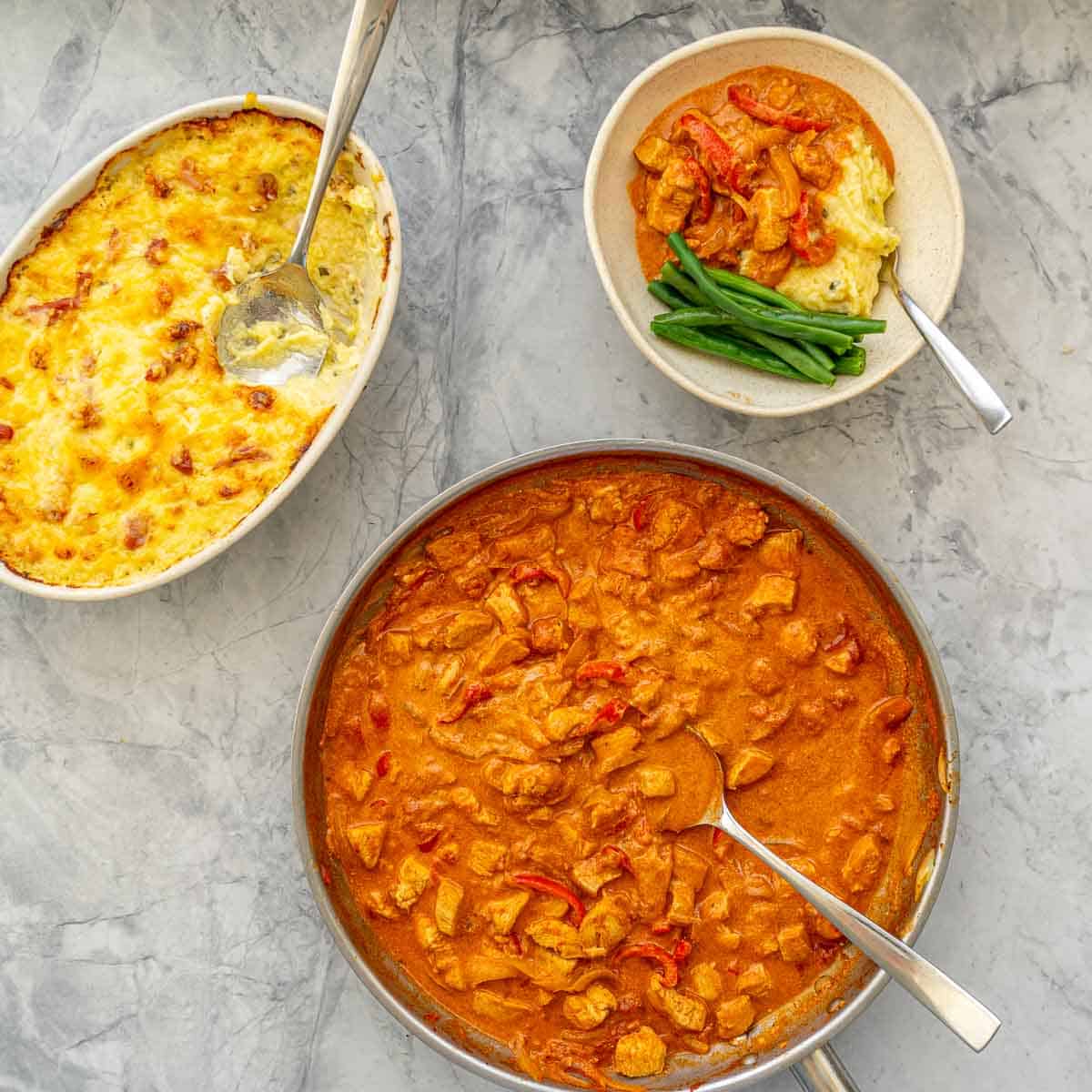 Dishes of Golden brown baked mashed potatoes and chicken Paprikash on the bench next to a serving of the Chicken Parprikash over a big spoonful of the-potatoes. anda side of beans
