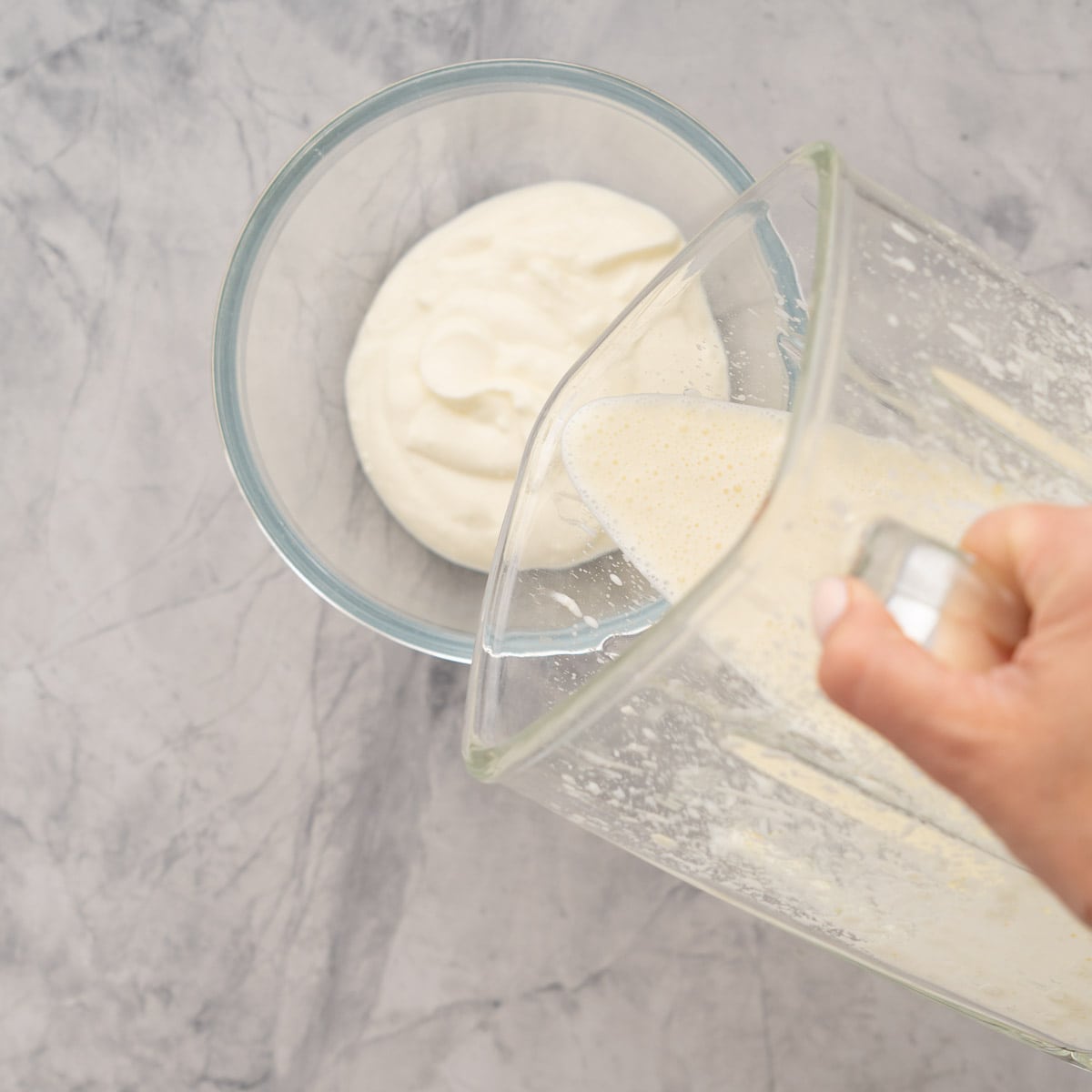 Feta and yoghurt mixture being poured from the blender into a second measure of yoghurt in a glass bowl.
