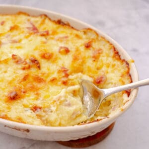 A ceramic serving dish of baked Mashed Potatoes with a large serving spoon resting on the side.