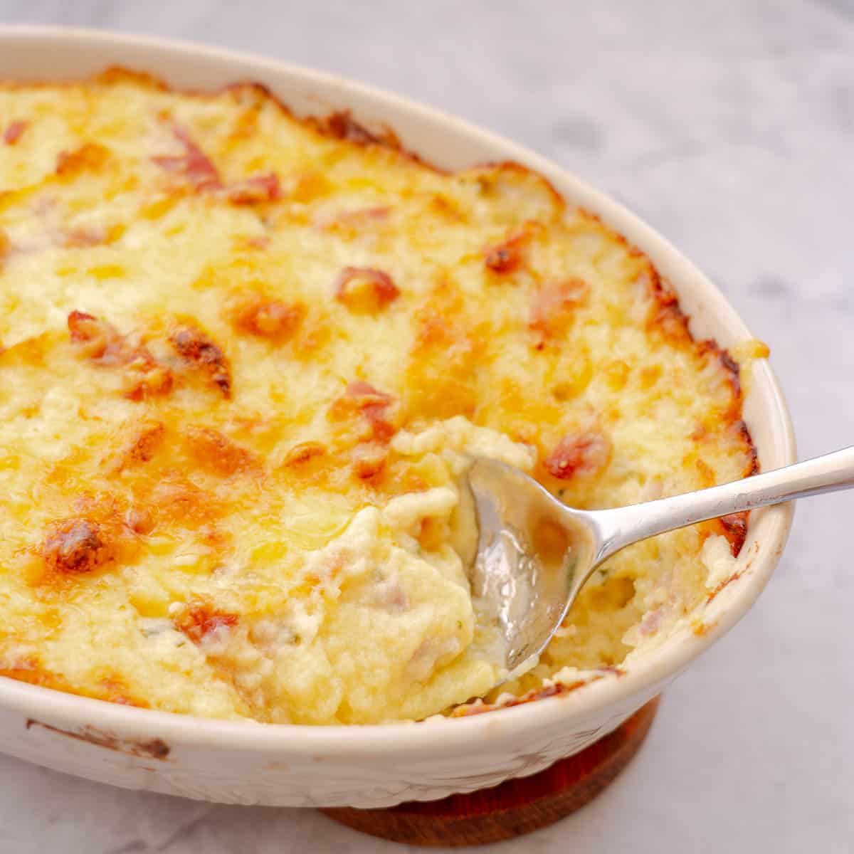 A ceramic serving dish of baked Mashed Potatoes with a large serving spoon resting on the side.