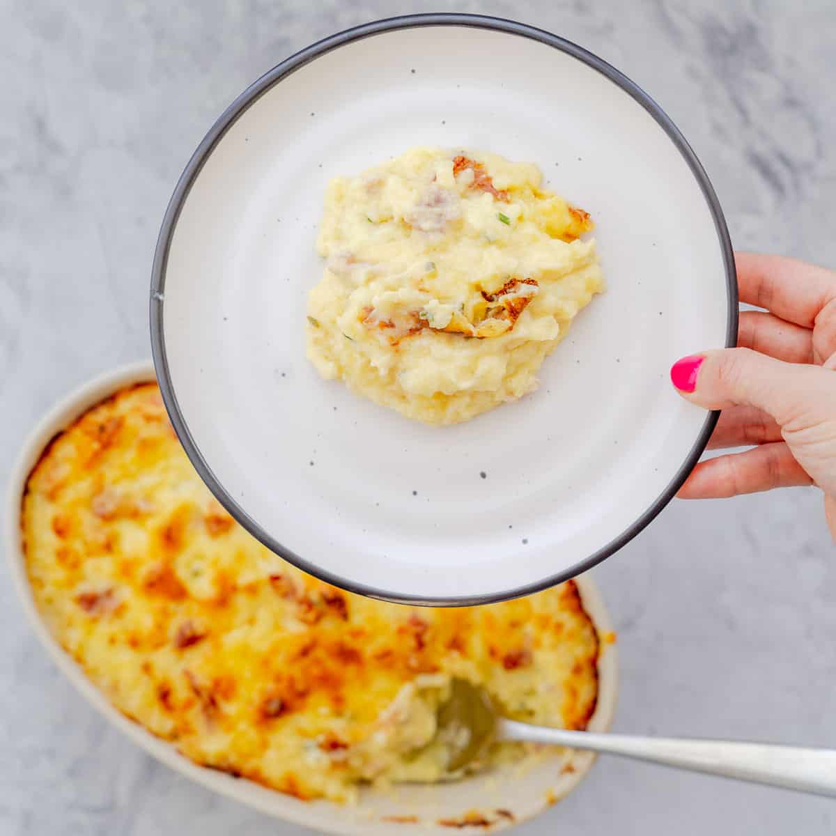 Golden brown Baked Mashed Potatoes resting the bench with a serving a plate held above.