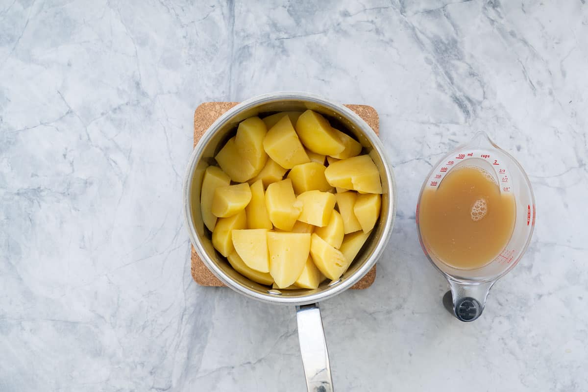 A saucepan of cooked cut ip potatoes sitting next to a jug of drained chicken broth.