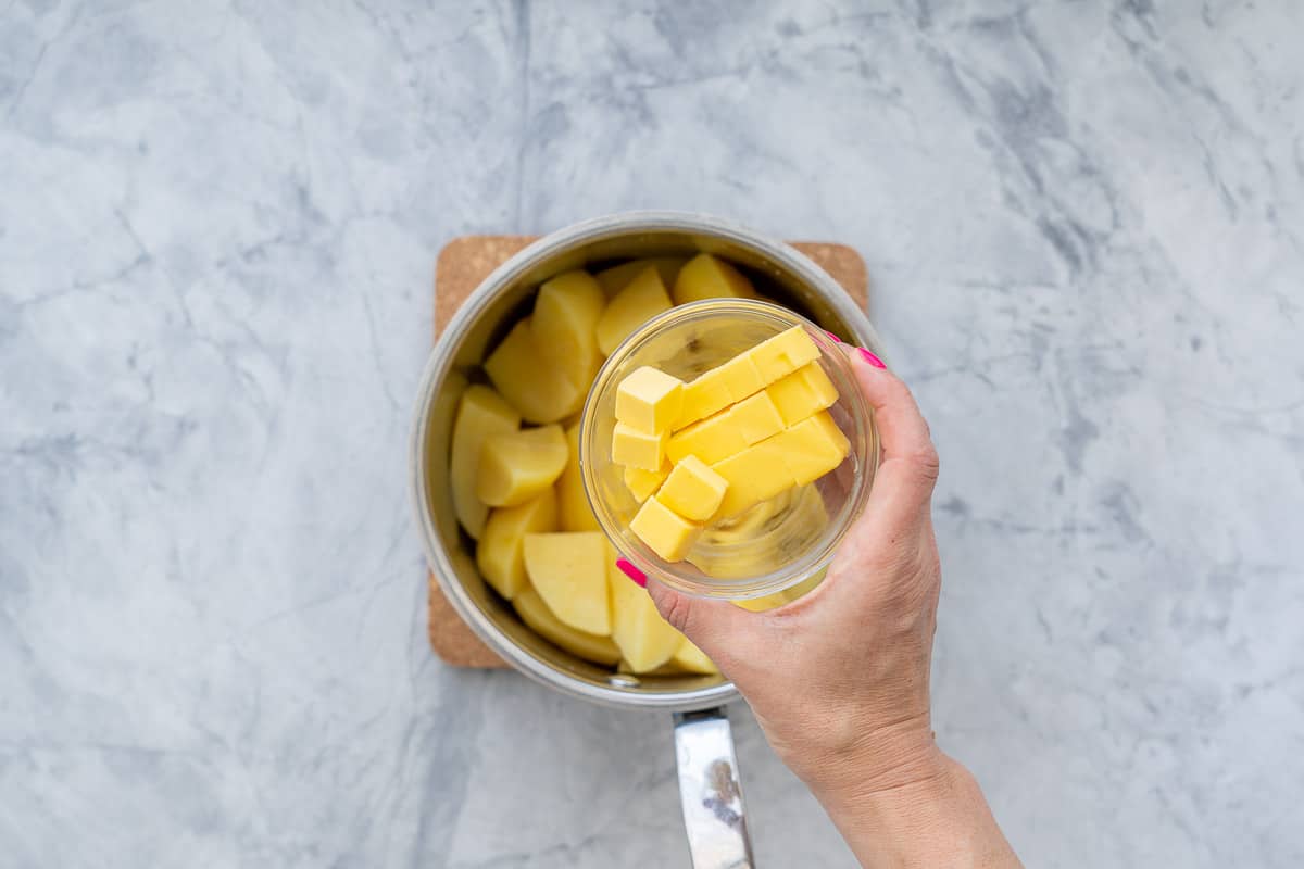 A hand holding a small glass ramekin of cubed butter above a saucepan of cooked cut up potatoes.