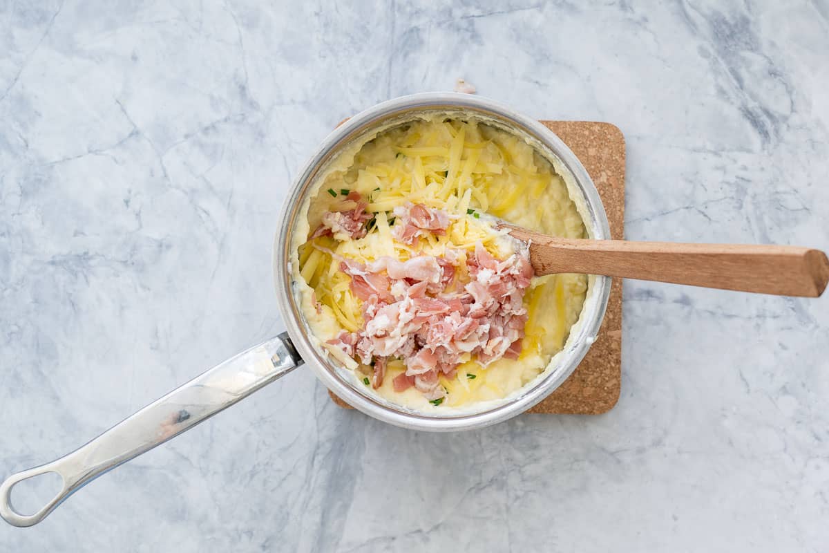 A saucepan of mashed potatoes with bacon added and a wooden spoon resting on the side.