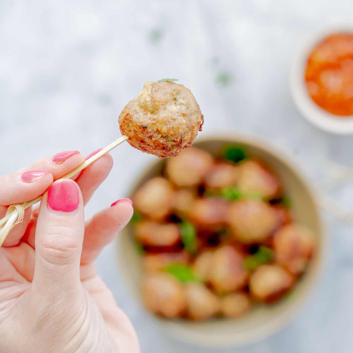 A bowl full of meatballs, with a hand holding up one meatball above them with a tooth pick.