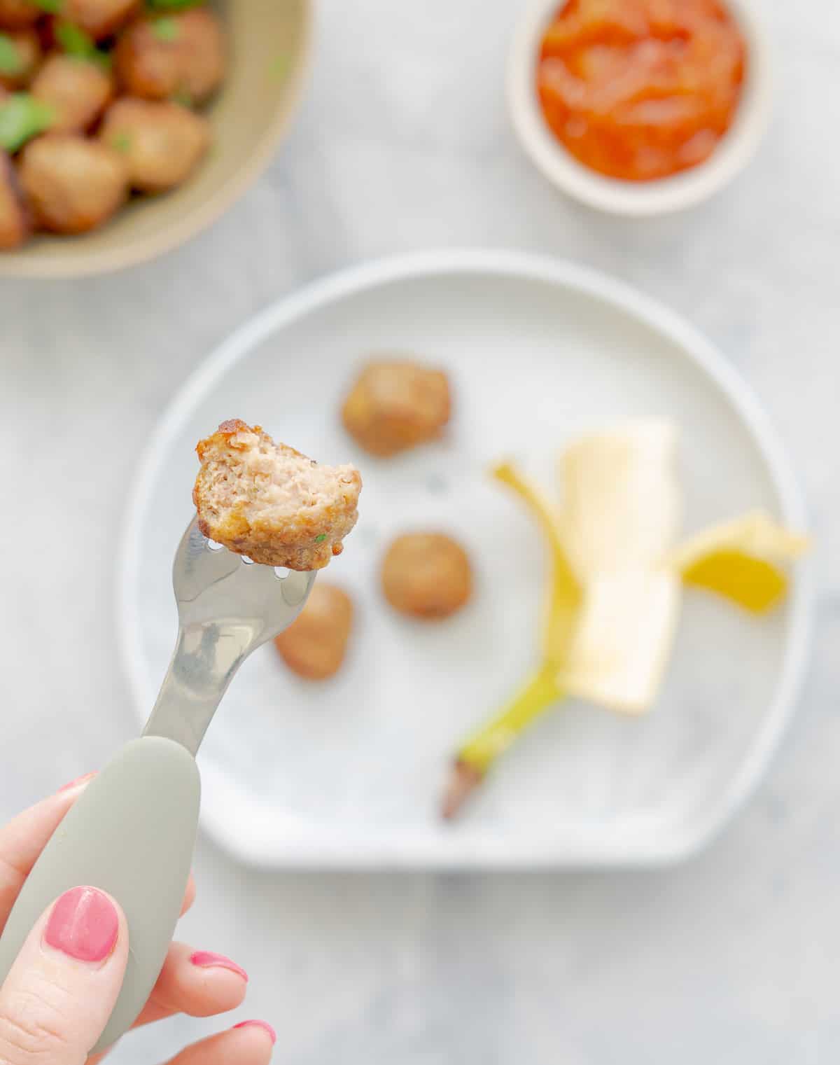 A serving of baked meatballs and half a banana on a silicon toddlers plate with a halved meatball held up by a toddlers fork above them.