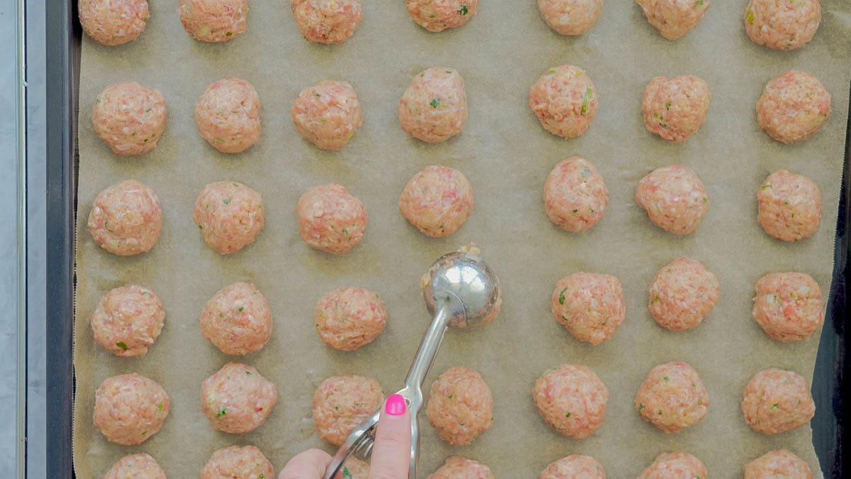 Scoops of golf sized meatball mixture with a cookie scooper placing the last one down on the lined baking tray.