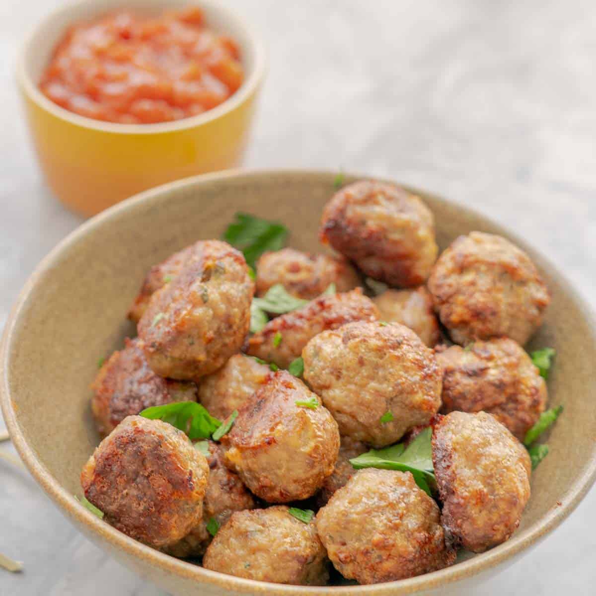 A ceramic serving bowl full of baked meatballs with a sprinkle of parsley next to a small ceramic serving dish of chutney