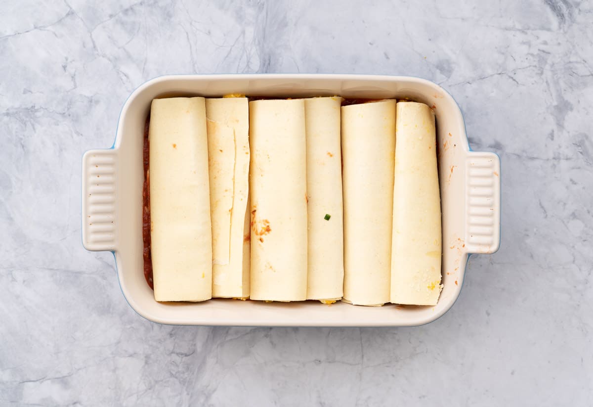 A baking dish sitting on a bench with 6 stuffed pasta rolls laid out in the bottom of it.