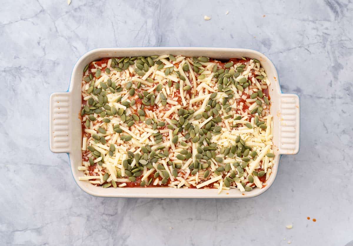 A baking dish sitting on a bench with stuffed pasta rolls topped with the pasta sauce, cheese and pempitas