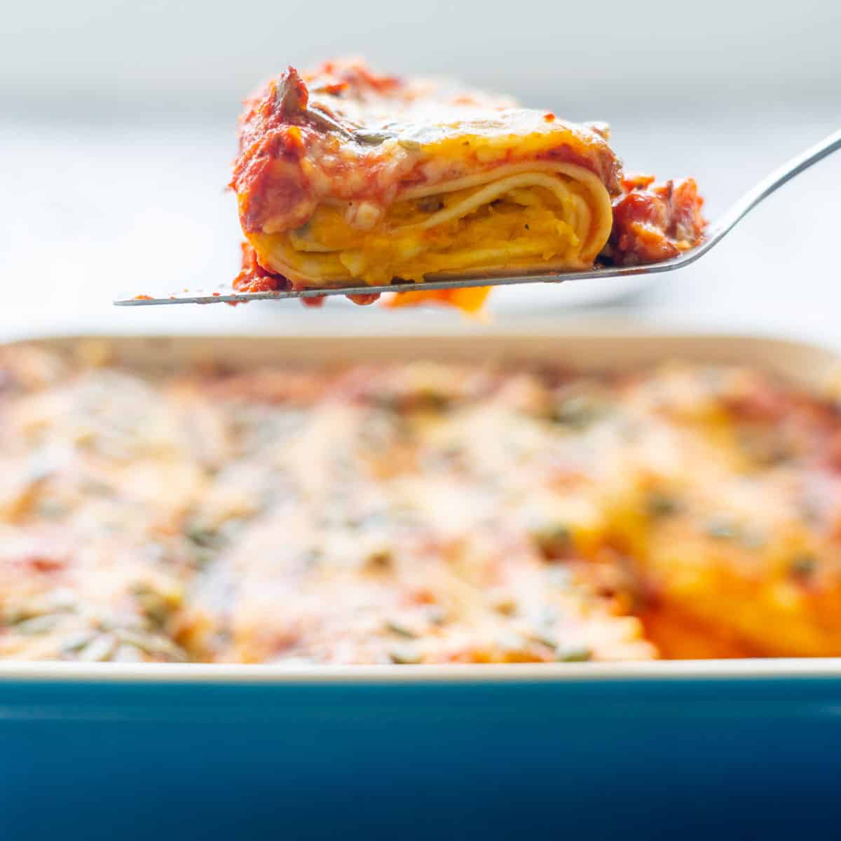 A baking dish of golden brown baked Butternut Squash Pasta cooling on a baking rack on the bench with a slice held above by a spatula
