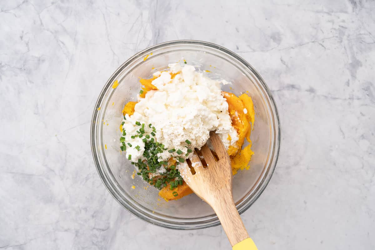 Roasted butternut, cottage cheese, chives and salt and pepper in a glass mixing bowl with a wooden spoon resting on the side.