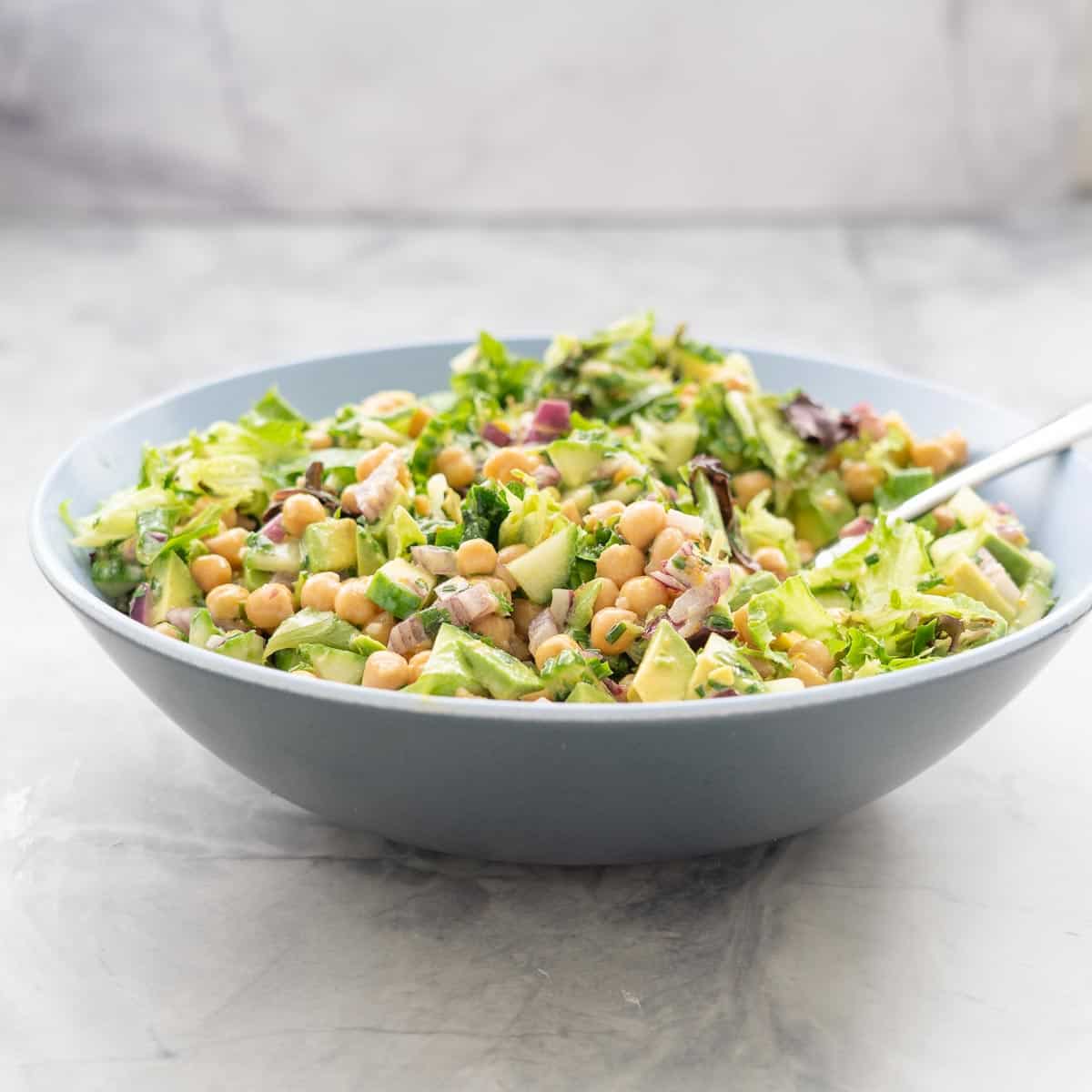 A salad made with chickpeas and avocados in a large blue ceramic bowl, chopped red onion, herbs and lettuce are also visible in the salad.