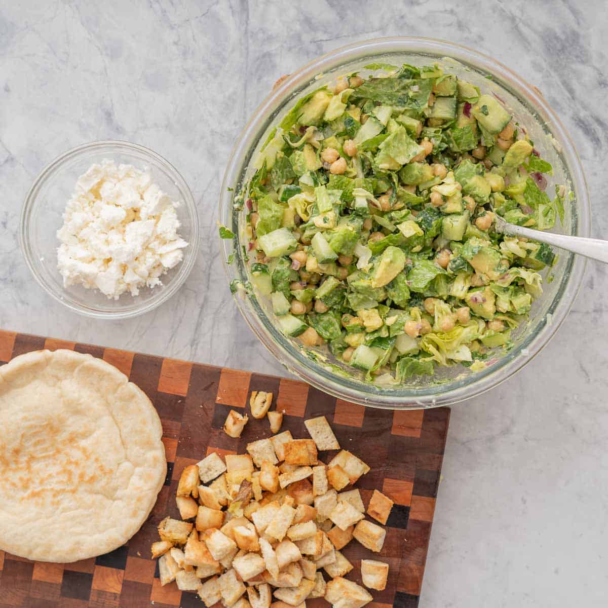 A large glass bowl of chickpea avocado salad on a bench with a small bowl crumbled feta and pita bread.