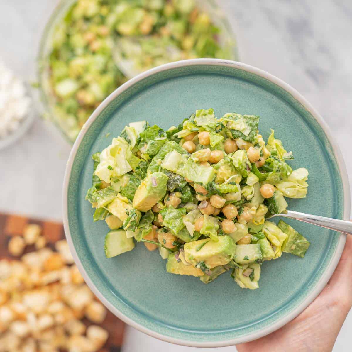A salad made with chickpeas and avocados on a turquoise plate being held up above a large bowl of salad.