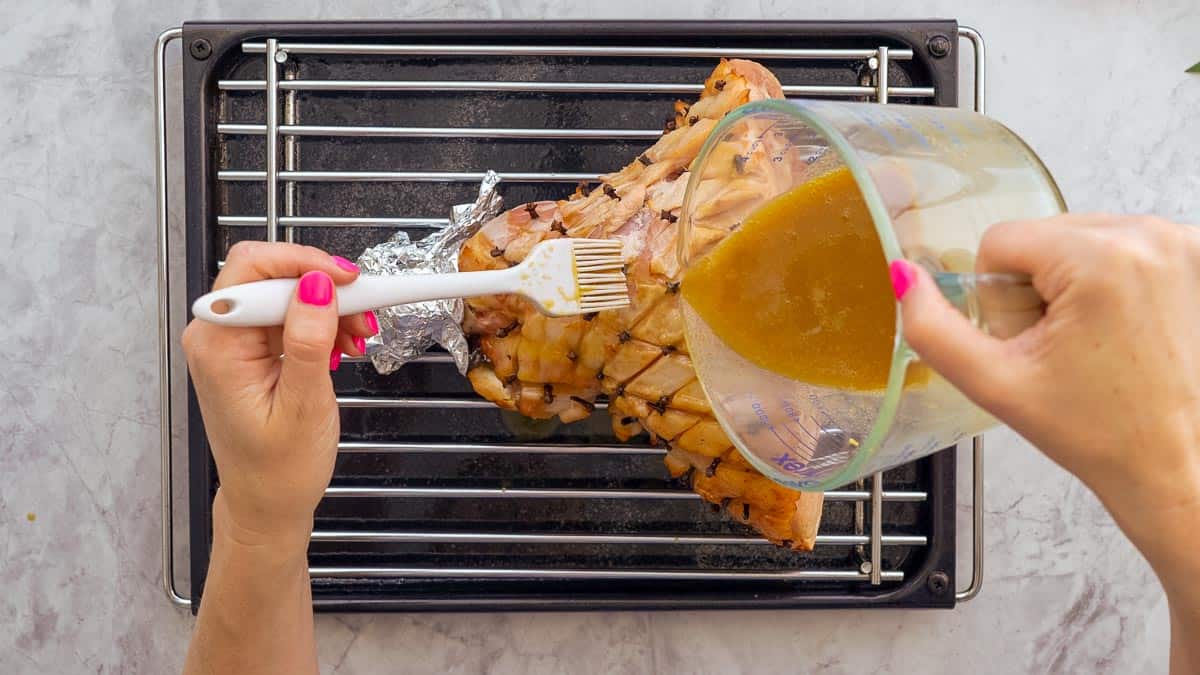 A jug of whisked glaze being poured over the top of the large cooked ham while a hand is brushing the glaze into the fat layer.
