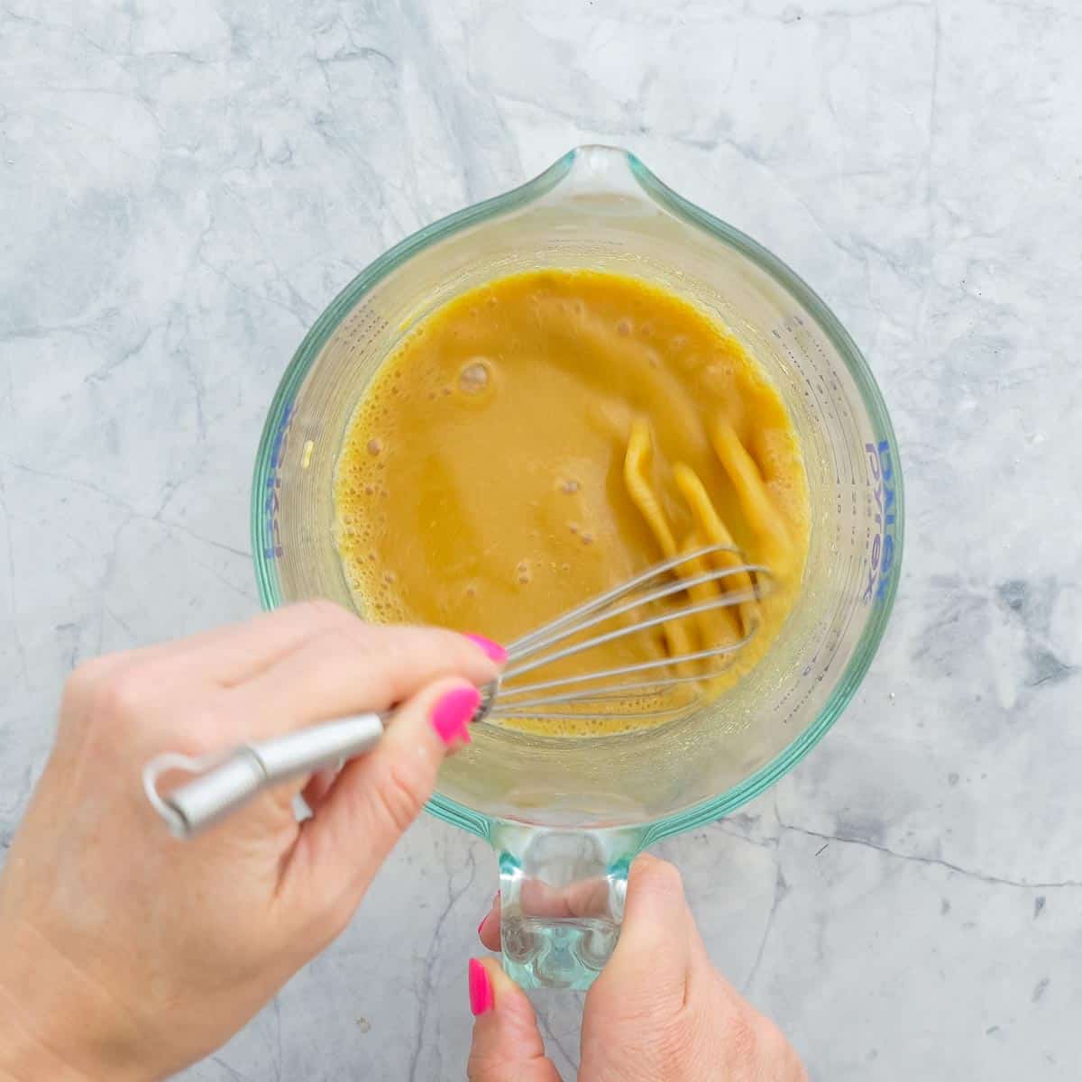 A small glass jar with hand whisking juice, brown sugar and mustard together on a bench