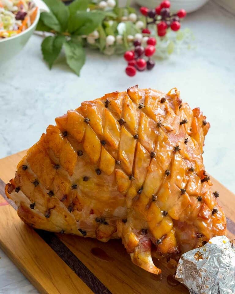 A cooked glazed ham with cloves pressed into the corners of diamond shaped cuts in the fat layer resting on a wooden chopping board on the bench with christmasy decorations in the background
