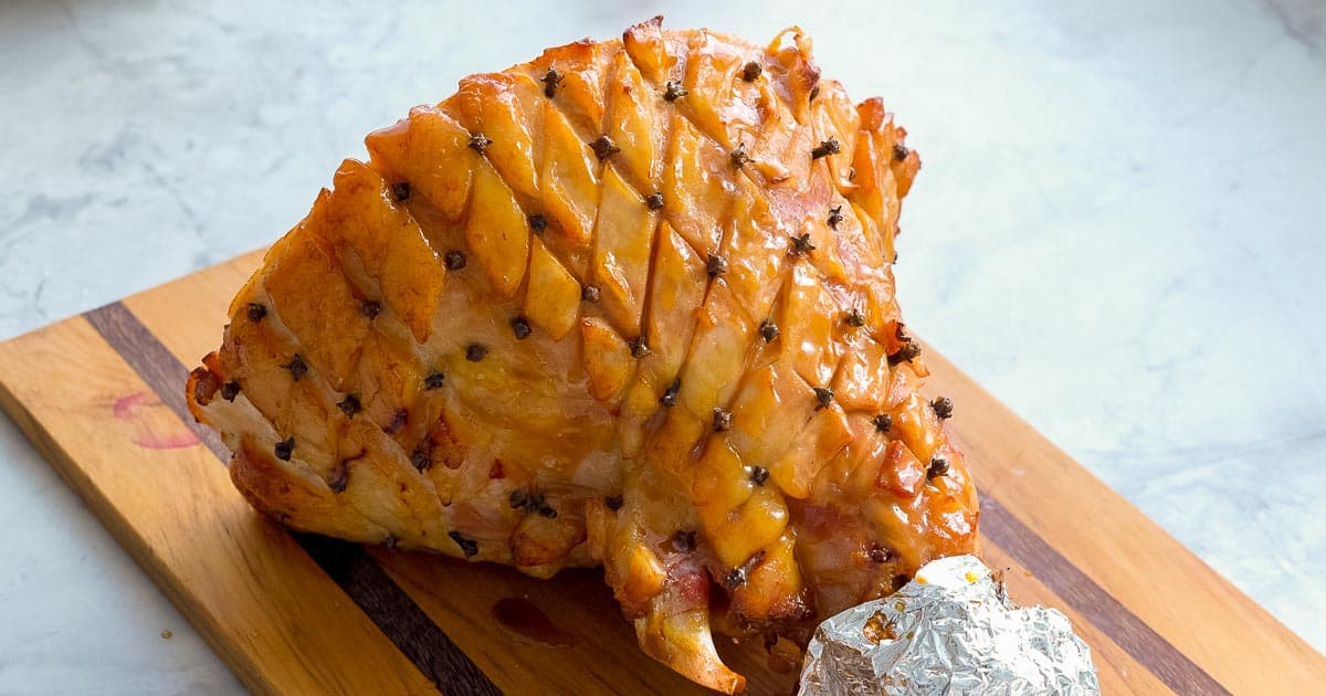 A cooked glazed ham with cloves pressed into the corners of diamond shaped cuts in the fat layer resting on a wooden chopping board on the