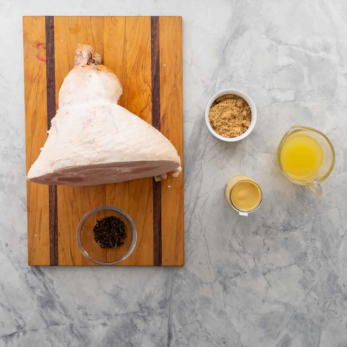 A large uncooked ham shoulder resting oil a wooden chopping board next to all the other ingredients to make the glaze