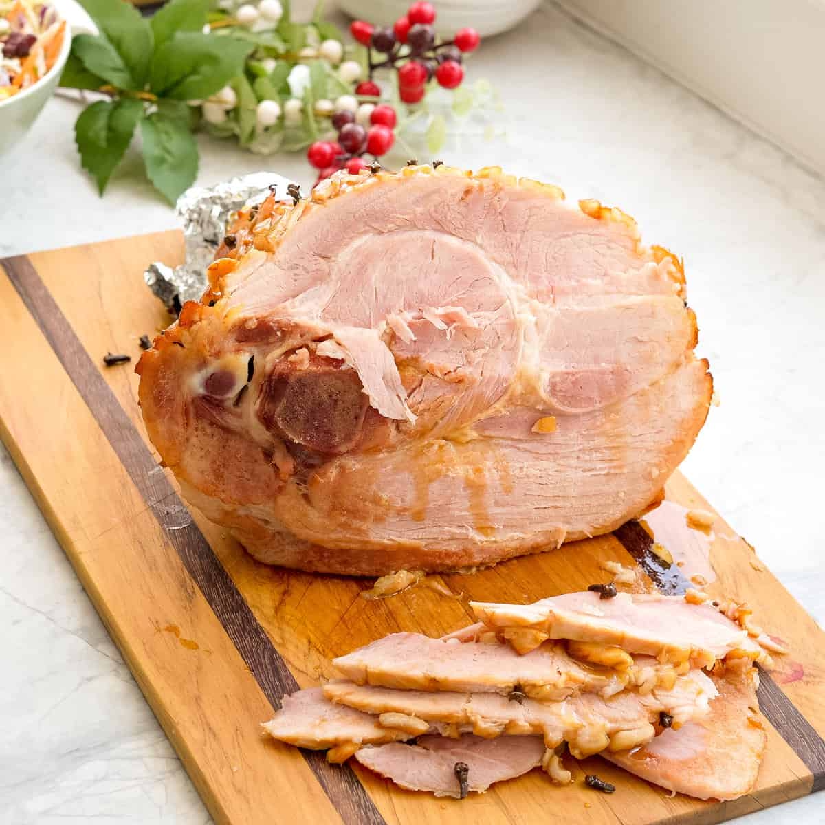 A large cooked ham resting on a wooden chopping board on the bench with multiple slices of ham beside it.