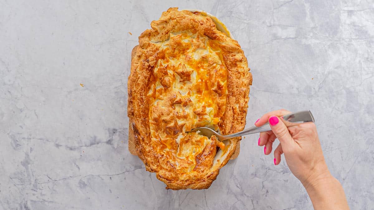 Golden brown baked ham pot pie resting on the bench with a hand taking a serving out with a large spoon