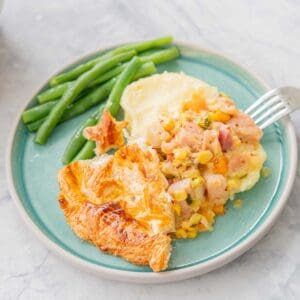 a serving of Ham Pot Pie with a side of blanched beans on a teal coloured ceramic plate and with a fork resting on the side, sitting the bench